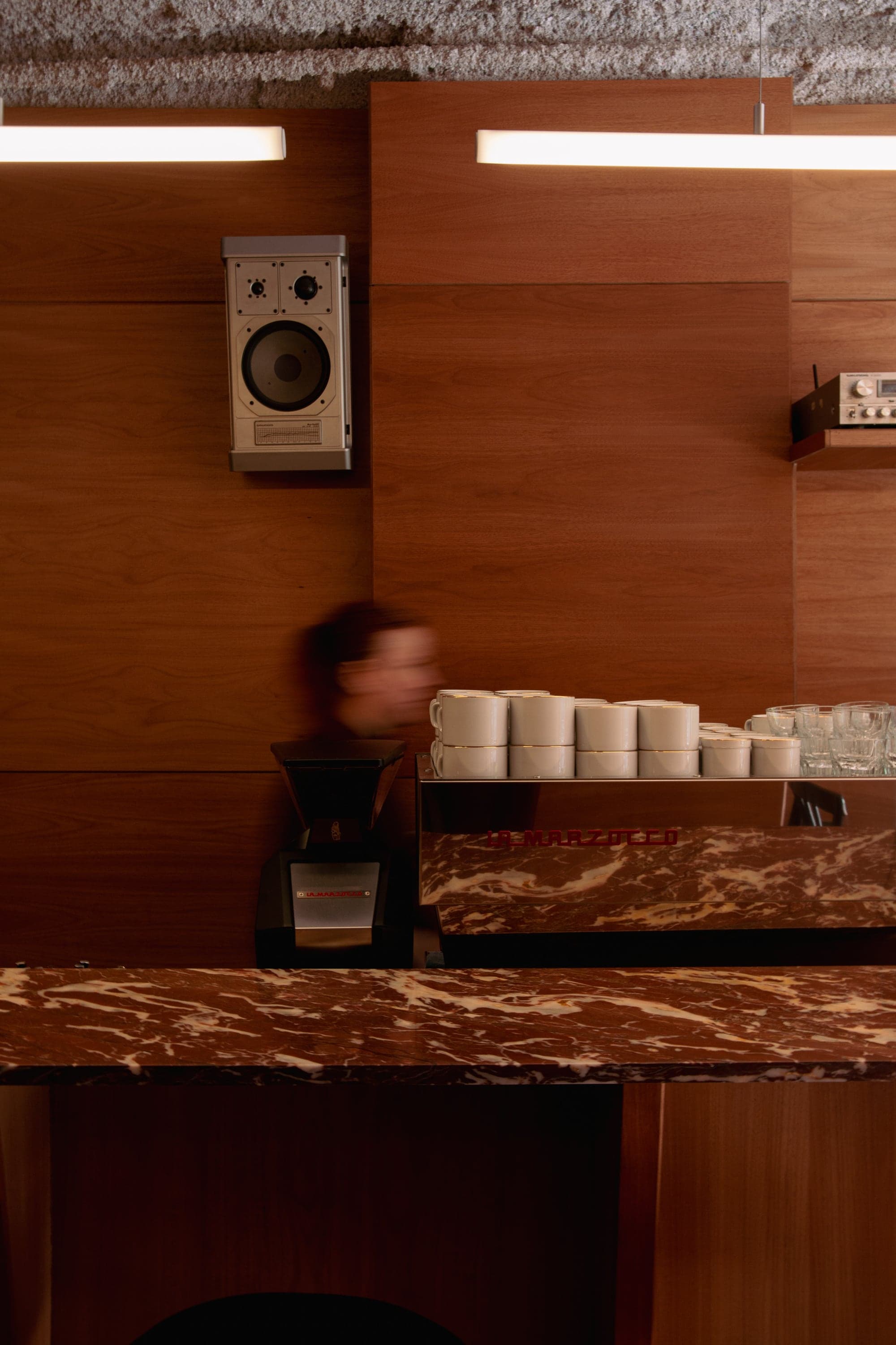 Detrás de la barra: paneles de madera, un parlante colgado en la pared, la máquina de espresso y tazas apiladas sobre el mármol rojo